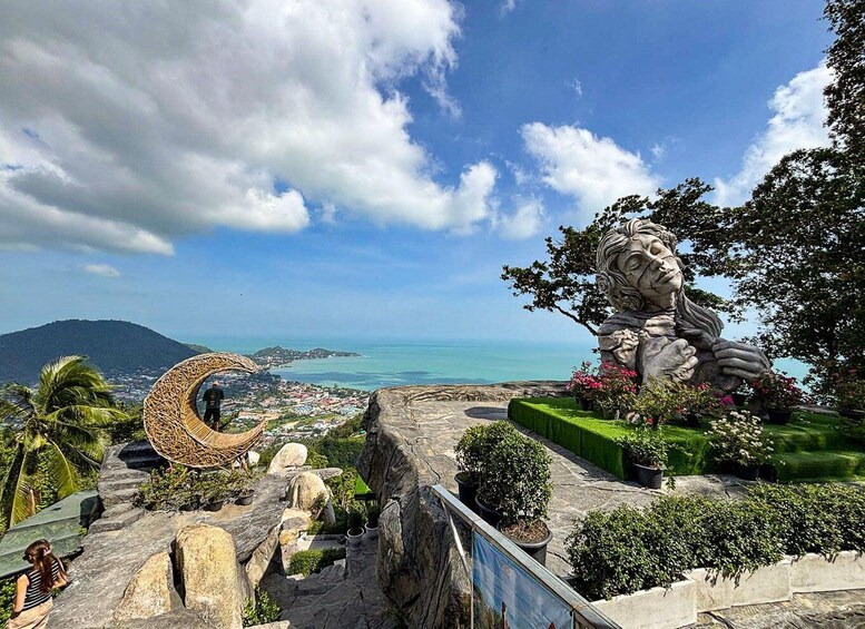 A scenic overlook with a stone sculpture, a circular woven structure, and a view of the coastline. 2