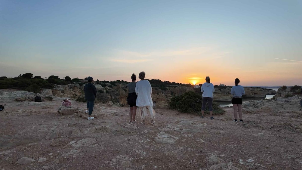 A group of people standing on rocky terrain, overlooking a sunset over a coastal landscape. 4