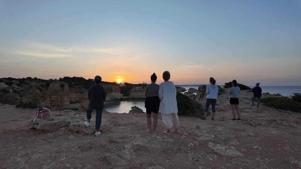 A group of people standing on a rocky cliff overlooking a sunset over a body of water. 5