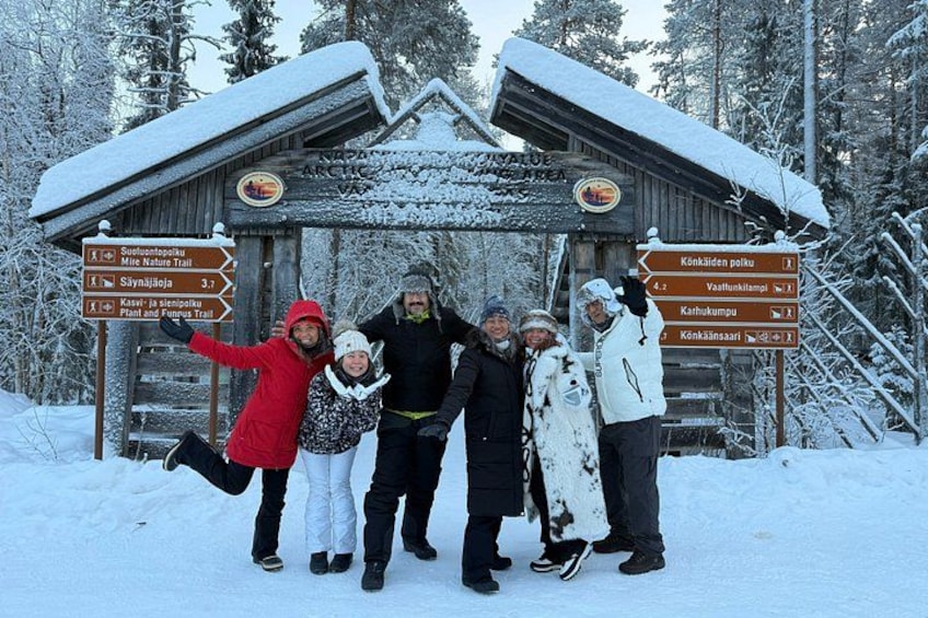 Eine Gruppe von Personen posiert vor einer Holzhütte mit einem Wegweiser, der verschiedene Wanderwege zeigt. 1