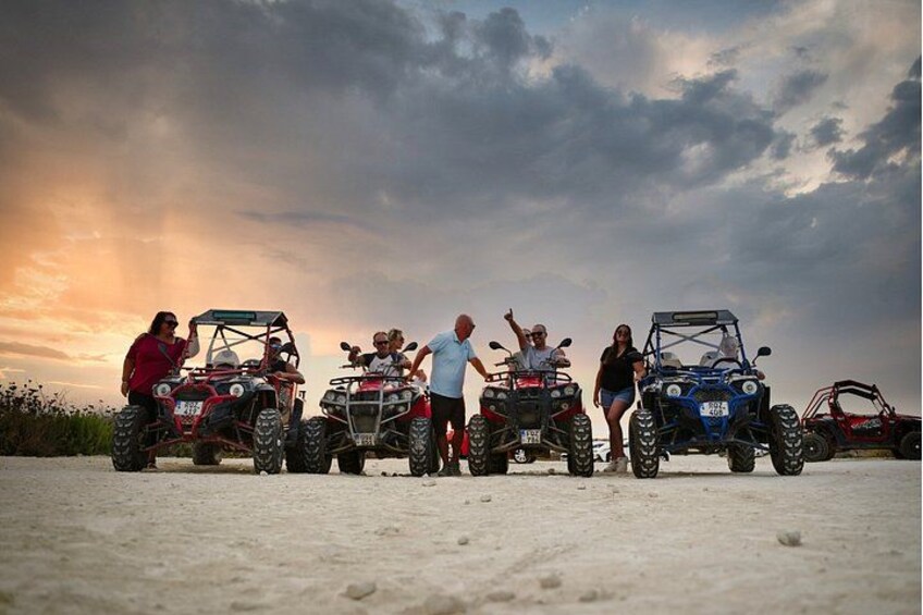 A group of people standing beside atv vehicles on a sandy surface with a cloudy sky in the background. 1