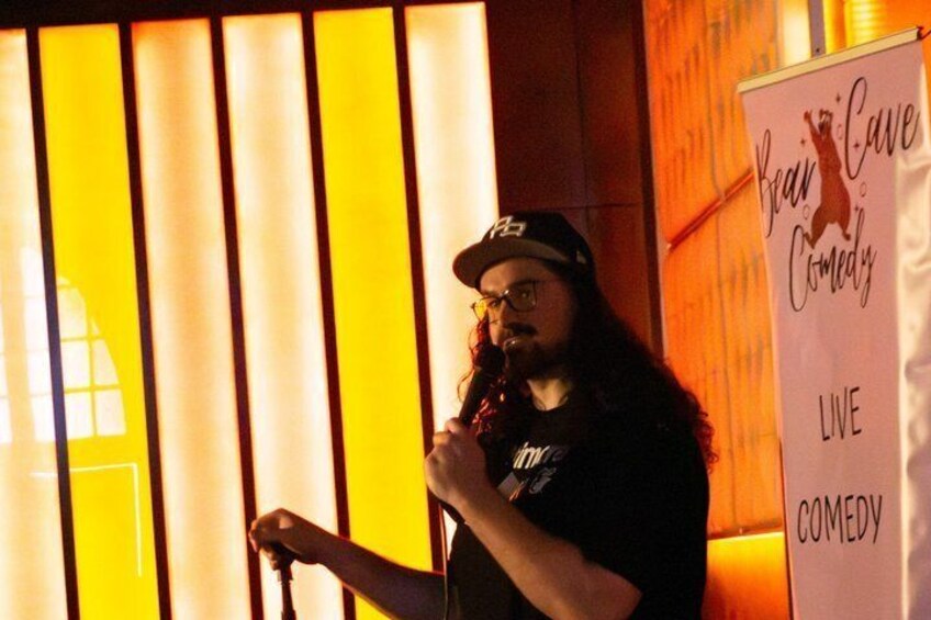 A person holding a microphone in front of a sign that reads 'Bear Cave Comedy Live Comedy.' 4