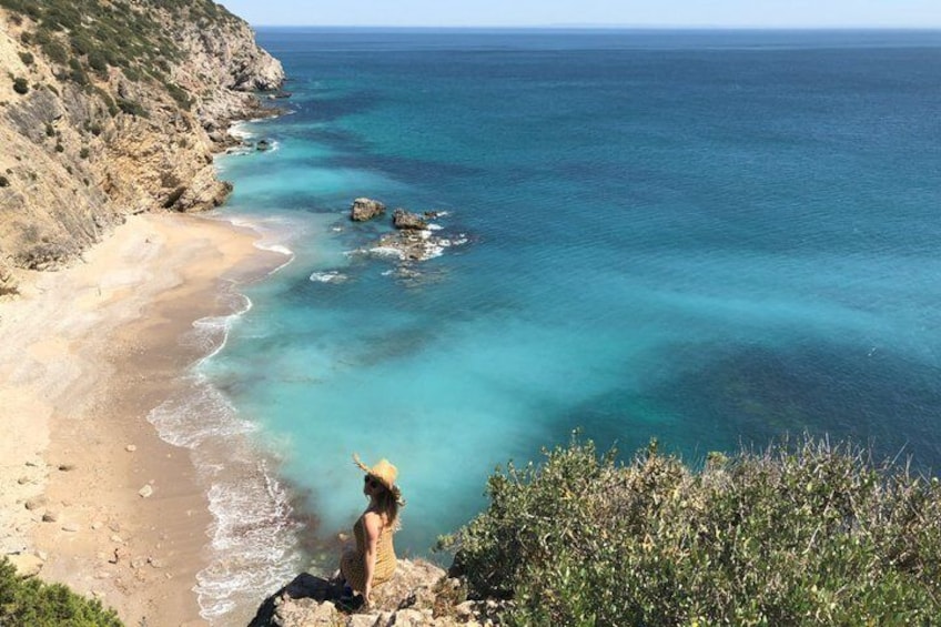 Un paysage côtier avec une mer bleue et limpide, une plage de sable, des falaises rocheuses et une personne debout sur une falaise, surplombant l’eau. 1