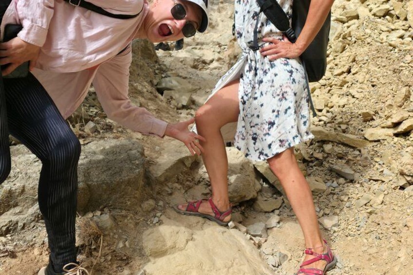 Une personne portant une chemise rose et un pantalon rayé aide une autre personne, vêtue d’une robe à motifs floraux et de sandales, à se déplacer sur un terrain rocheux. 2
