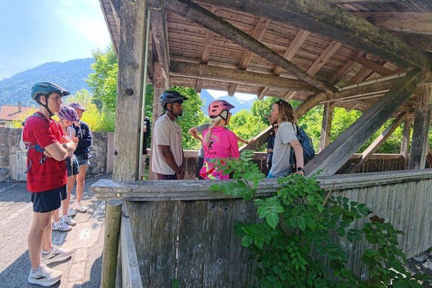 Un grupo de personas se refugian bajo una estructura de madera, algunas con casco, con follaje y montañas al fondo. 2