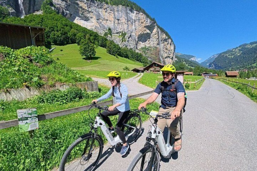 Two cyclists on a paved path with greenery and a mountain backdrop. 2