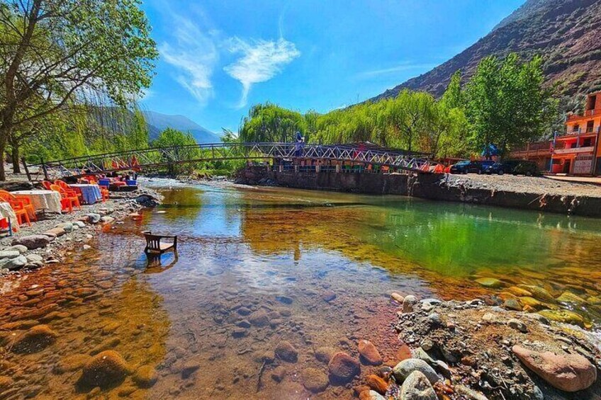 A clear river with rocks and a wooden chair in the water, surrounded by trees and a bridge. 2