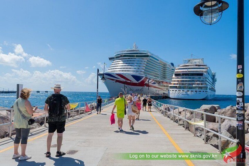 A pier with people walking, a large cruise ship, and a smaller ship in the background. 3