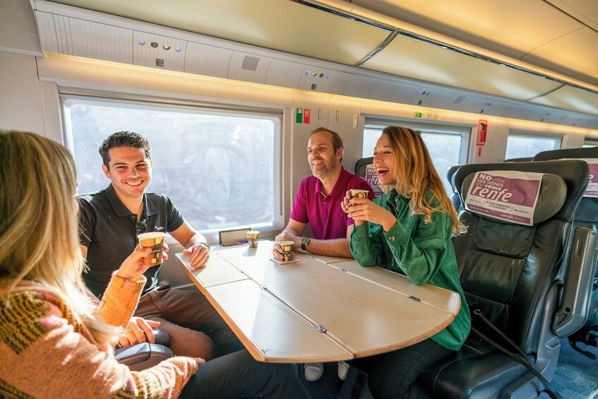 Four people seated at a table inside a train carriage, each holding a cup, with a sign on the seat reading 'No todos los trenes son iguales.' 3