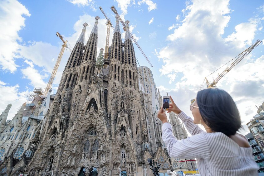 A person taking a photo of a building with intricate architecture and construction cranes in the background. 1