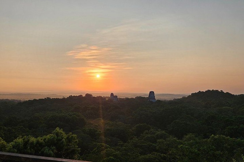 Un atardecer sobre un paisaje boscoso con una colina distante y una pequeña construcción. 3