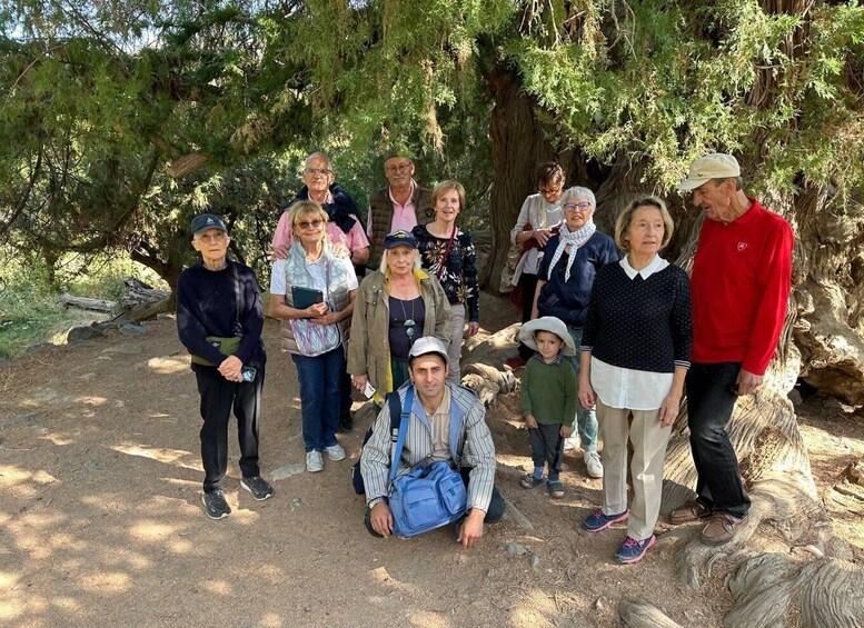 A group of people gathered outdoors, some standing and some sitting, with trees and a rocky ground in the background. 4