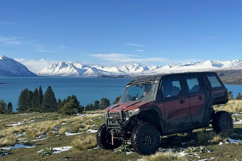 A red off-road vehicle parked on a grassy field with snow patches, a lake, and snow-capped mountains in the background. 2