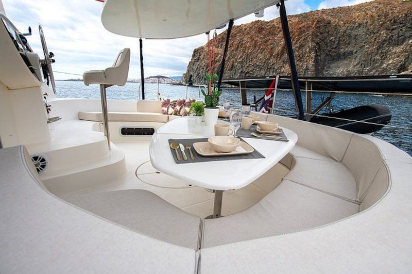 A boat's deck with a dining table set for two, surrounded by white cushioned seating. 2