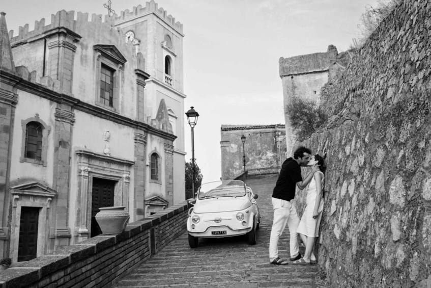 A historic building with a clock tower, a vintage car parked in front, and a couple kissing on a stone wall. 2