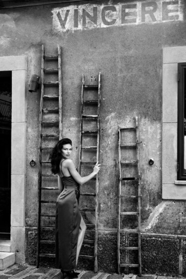 A woman standing next to a building with two ladders leaning against it and the word 'VINCERE' painted on the wall. 5