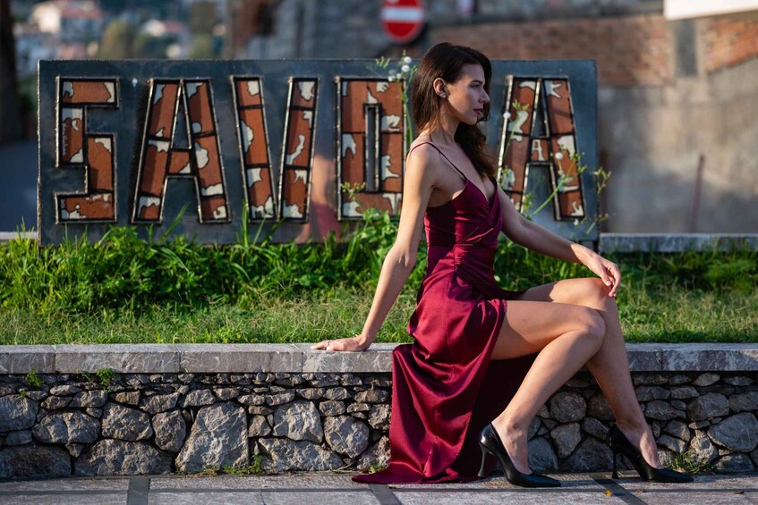 A woman in a red dress sitting on a stone wall with a sign behind her that reads 'SAVIA.' 1