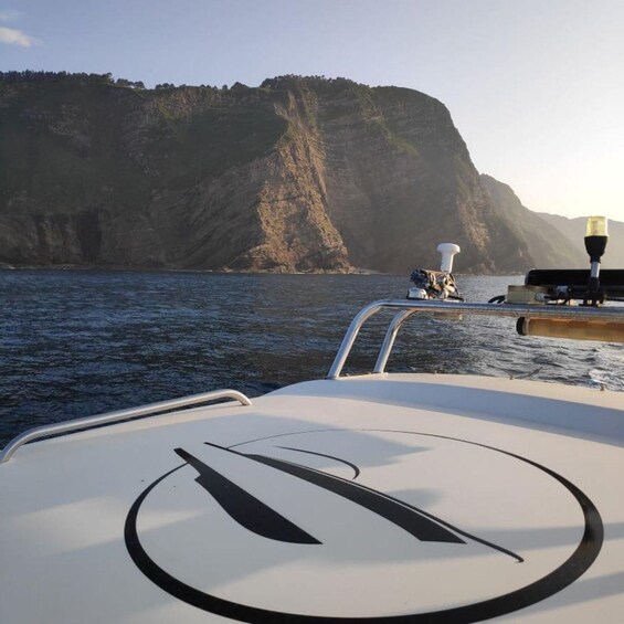 A boat's deck with a logo, a rocky cliff, and calm water. 3