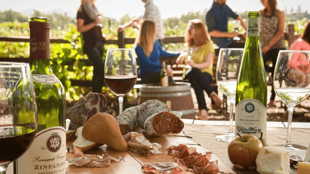 A table with wine bottles, glasses, cheese, and cured meats at an outdoor gathering. 4