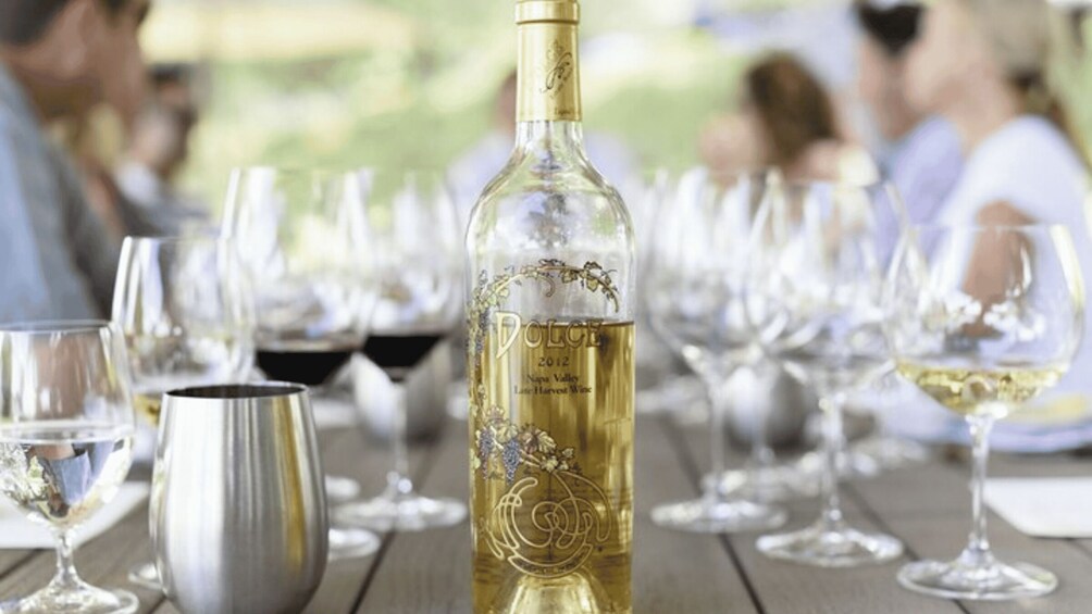 A bottle of Dolce 2012 Napa Valley Late Harvest Wine on a wooden table surrounded by wine glasses. 1