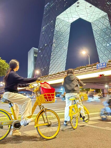 Two people riding bicycles with baskets, passing by a modern building with a distinctive geometric facade. 2