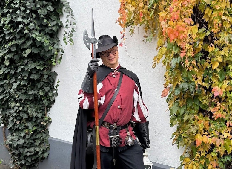 A person in a red and black costume holding a sword, standing in front of a wall with ivy and autumn leaves. 1