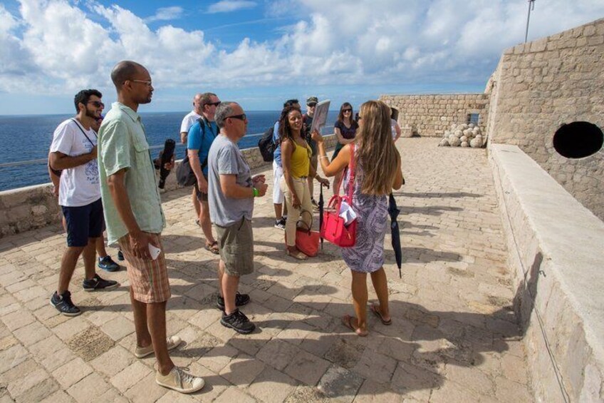 Eine Gruppe von Menschen steht auf einer steinbelagten Fläche am Meer, über ihnen ein klarer Himmel. 1