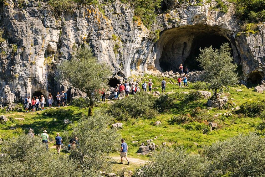 A group of people hiking near a rocky cliff with a cave entrance. 3