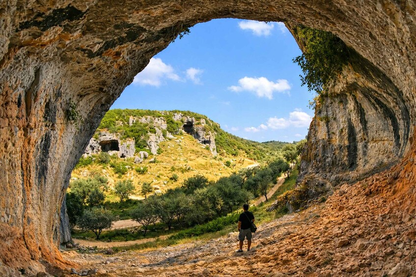A person standing in a rocky landscape with a cave entrance and a hill in the background. 1