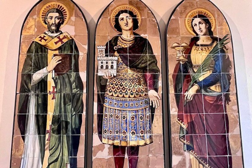 A stained glass window with three figures, each adorned with halos and holding symbolic objects. 5