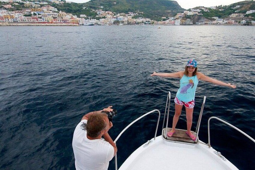 A person standing on a boat's deck with arms outstretched, facing a body of water with a coastal town in the background. 5