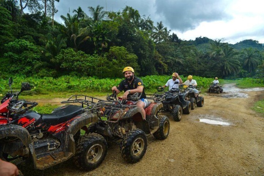 Een groep mensen op quads op een onverharde weg, met weelderige begroeiing op de achtergrond. 1