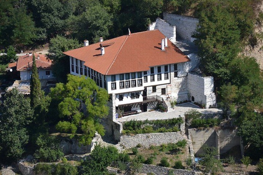 Un edificio grande y blanco con techo rojo, situado en una ladera rocosa rodeada de árboles. 1