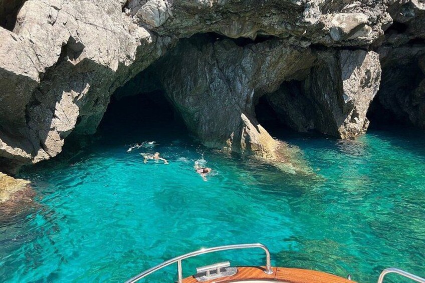 A clear blue sea with a rocky cave entrance, a boat in the foreground, and people swimming. 2