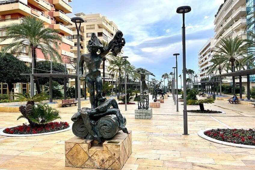 Una estatua de un hombre con una lira, rodeada de palmeras y bancos en una plaza con edificios al fondo. 5
