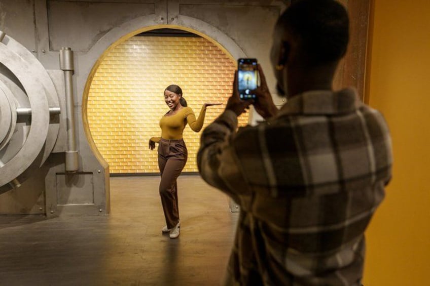 A person taking a photo of another person in a room with a brick wall and a large circular structure. 2
