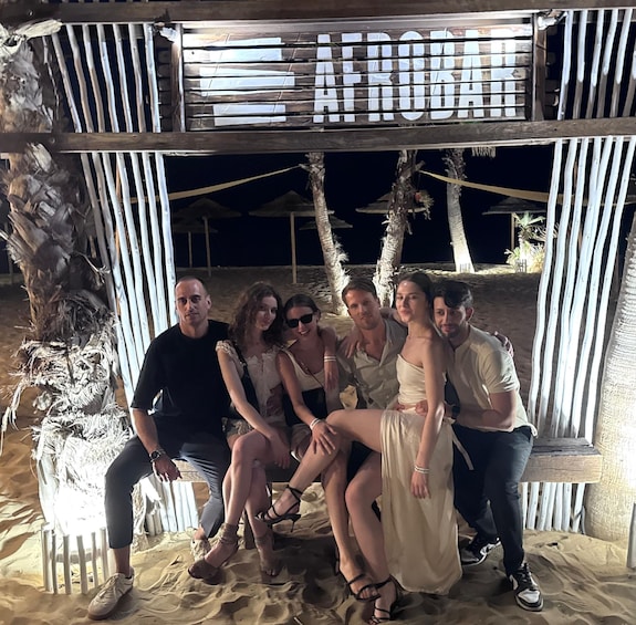 A group of people seated under a wooden structure with the word 'ATROCHE' on it, at a beach with palm trees and thatched umbrellas. 2