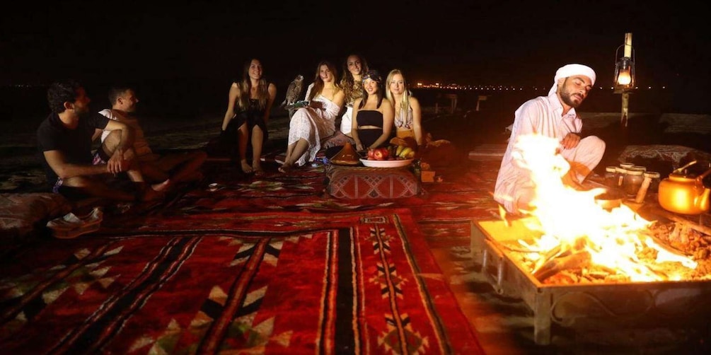 A group of people gathered around a fire on a patterned carpet, with a lantern in the background. 1