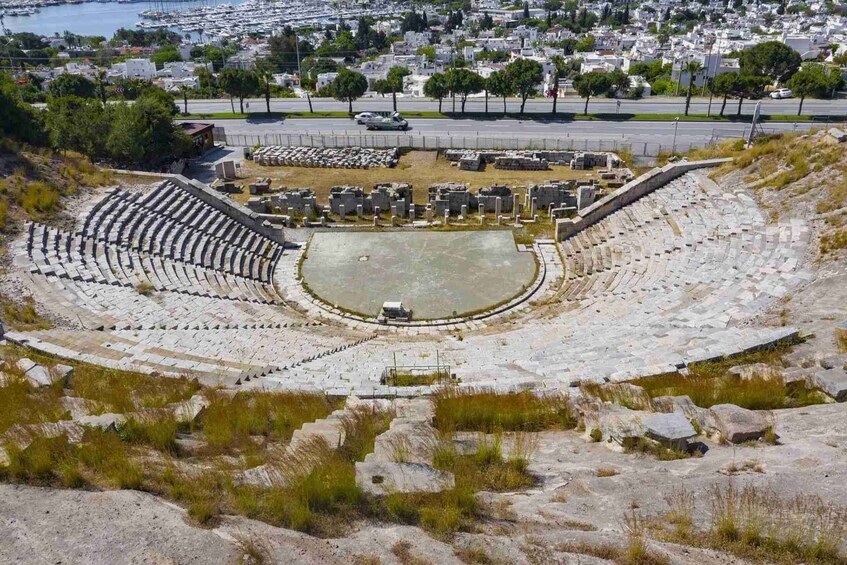 An ancient amphitheater with stone seating and a central stage, surrounded by ruins and vegetation. 5
