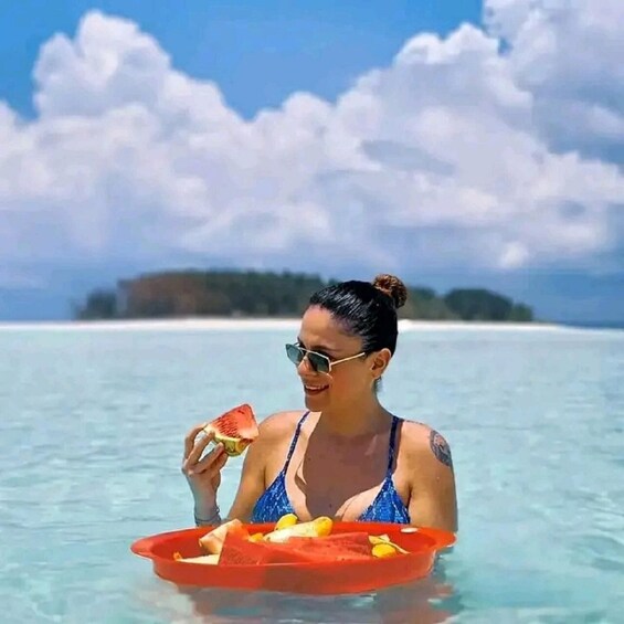 A person in a blue swimsuit holding a tray with fruits and a slice of watermelon in clear tropical waters. 1