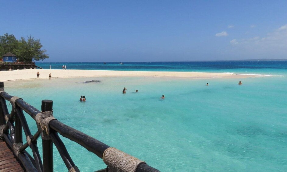 A beach with clear turquoise water, a wooden walkway, and a few people swimming. 4