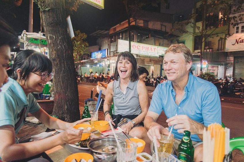 Un grupo de personas cenando al aire libre de noche, con mesas, comida y bebidas. 3