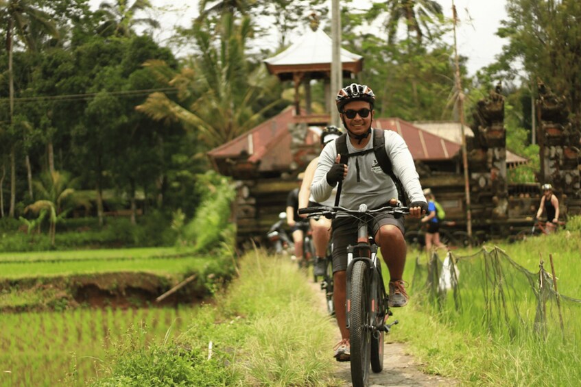 A person riding a bicycle on a rural path surrounded by lush greenery and traditional architecture. 3