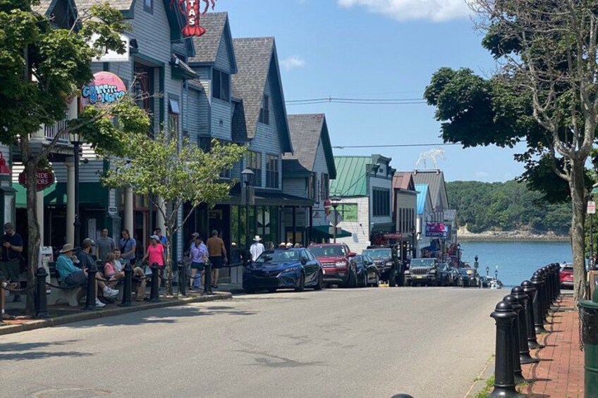 A waterfront street with shops, parked cars, and people sitting on benches. 2