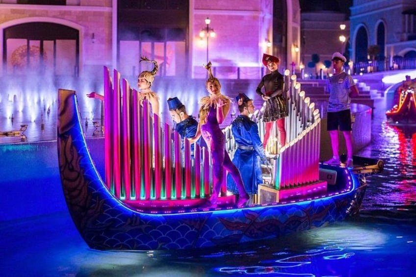 Un bote con gente a bordo, iluminado por luces de colores, sobre un cuerpo de agua con fuentes al fondo. 1
