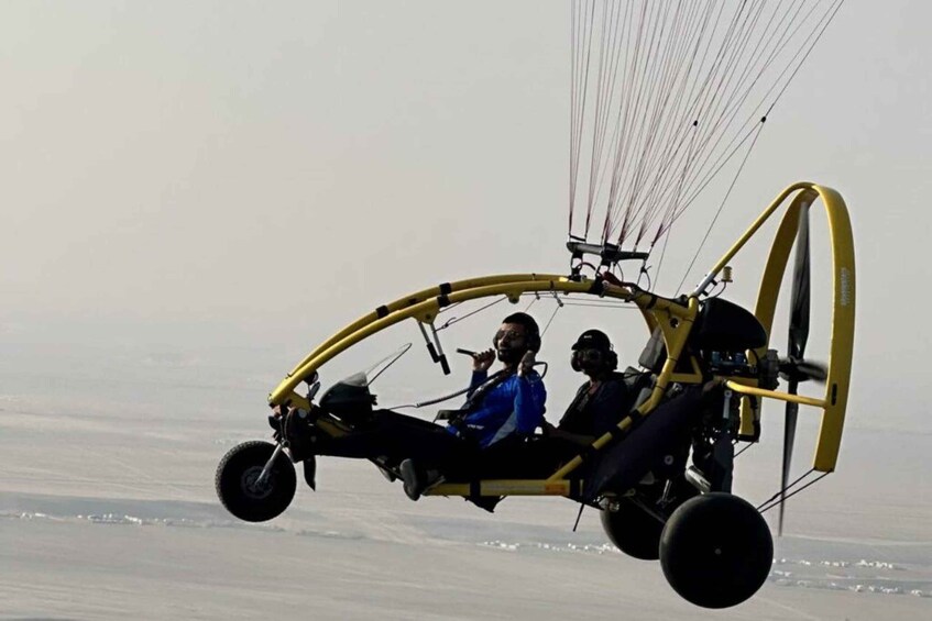 A yellow hang glider with two passengers in mid-flight over a flat landscape. 2