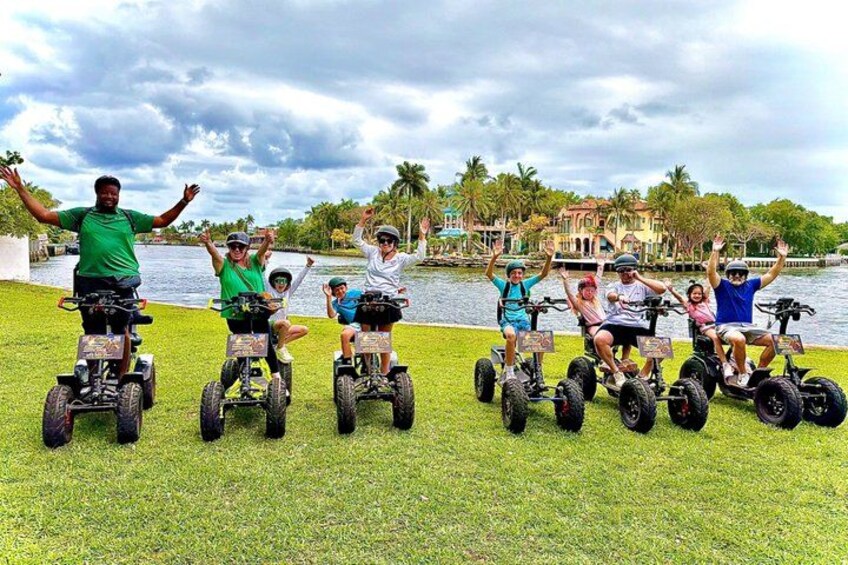 A group of people on Segway ATVs in a grassy area with a lake and buildings in the background. 1