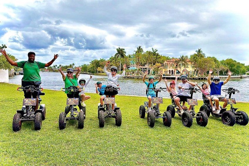 People on Segways in a grassy area with a lake and buildings in the background. 1