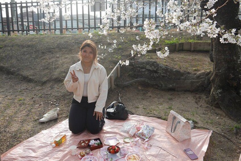 A person sitting on a picnic blanket with food and a bag, surrounded by trees and a fence. 3