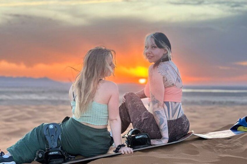 Zwei Personen sitzen auf einer Decke am Strand, im Hintergrund ein Sonnenuntergang. 4